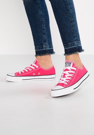 Nohy v jasně růžových nízkých teniskách Converse s bílými tkaničkami a podrážkami, spojené s tmavě modrými úzkými džíny se třepeným lemem.