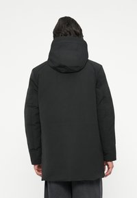 Melns, izolēts parka mētelis ar kapuci, demonstrējot gludu tekstūru, vienkāršu dizainu un garu siluetu. Minimalistiska stila ar redzamu aparatūru.