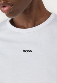 Bílý tričko s kulatým výstřihem a černě vyšitým logem "BOSS" na hrudi. Látka vypadá hladce a je lehká.