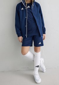 Laivastonsinen urheiluasu, johon kuuluu takki ja paita, yhdistettynä shortsseihin ja polviasteisiin valkoisiin sukkuihin. Huomattava Adidas-logo valkoisena vaatteissa ja kengissä.