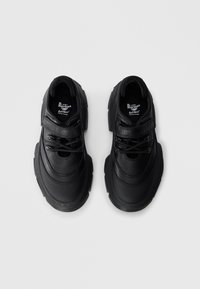 O pereche de pantofi negri Dr. Martens SoftWair din piele, cu tălpi groase, prinsi cu sireturi și o curea cu velcro, arătați dintr-o parte de sus pe un fond alb.