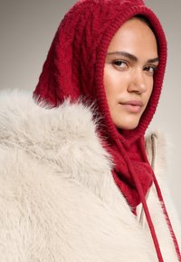 Rdeča pletena kapa s teksturiranim vzorcem v kombinaciji s kremasto ovratnico iz umetnega krzna. Jakna ima zadrgo in mehke, puhasto poudarjene detajle.