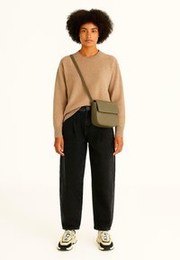 Beige gebreide trui met hoge hals, zwarte losse jeans en een olijfgroene gewatteerde crossbody tas. Sneakers met witte en zwarte accenten.