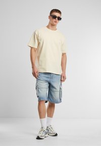 Šviesiai smėlinės spalvos medvilnės t-shirt, laisvas kirpimas, derinama su šviesiai mėlynomis džinsinėmis krovinių šortais, turinčiais šonines kišenes. Aksesuarai: juodi saulės akiniai ir sportbačiai.