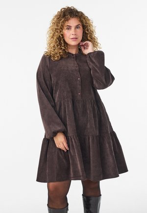 Mujer de cabello rizado que lleva un vestido suelto de manga larga en tonos marrón oscuro, medias negras y botas negras hasta la rodilla sobre un fondo liso.
