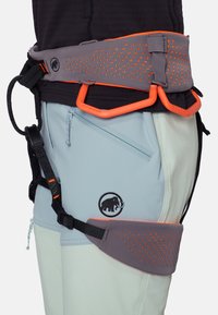 Klimtuig met grijze gewatteerde taille- en beenlussen, oranje uitrustingslussen en zwarte verstelbare banden, gedragen over lichtgekleurde broek.