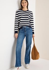 Gestreifter Pullover in Marineblau und Weiß, blaue Jeans mit lockerem Schnitt, weiße Sneaker mit roten Akzenten und eine beigefarbene Wildleder-Schultertasche.