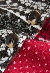 Nertos megztinis su tamsiai pilku ir baltu geometriniu raštu, dideliais rudais mygtukais ir raudona dalimi su mažomis baltomis dėmėmis.