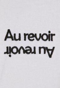 Balts audums ar melnu izšūtu tekstu, kas lasa "Au revoir" un "à tout à l'heure", attēlots otrādi. Gluda tekstūra, vienkāršs dizains.