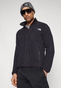 Svart fleecejakke med glidelåslommer, høy krage og The North Face-logo. Båret med en grå lue og solbriller, som gir et strukturert utseende.