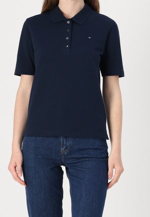 Vrouw draagt een marineblauw poloshirt met korte mouwen, vier knopen en een klein logo, gecombineerd met blauwe spijkerbroek.