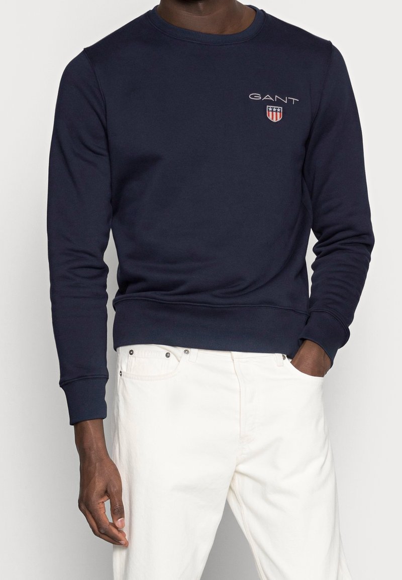 Sudadera navy hecha de algodón, con cuello redondo, puños y dobladillo acanalados, y un pequeño logo bordado en el pecho. Llevada sobre pantalones blancos.