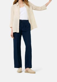 Kobieta ubrana w beżową koszulę zapinaną na guziki, noszącą białą bluzkę na ramiączkach, granatowe spodnie z troczkami i beżowe espadryle wsuwane, stojąca na tle białym.