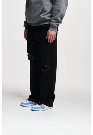 Persona che indossa una felpa grigia con cappuccio, jeans neri larghi strappati e sneakers bianche e blu, in piedi contro uno sfondo chiaro uniforme.