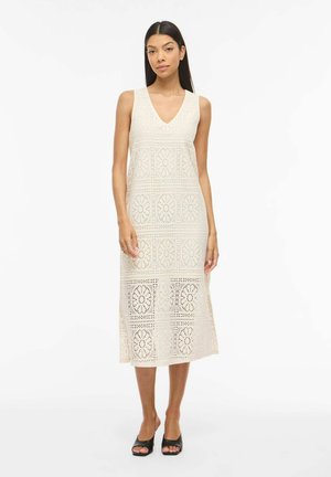 Femme portant une robe midi sans manches en crochet crème à motif floral et des talons ouverts noirs, debout sur fond blanc.