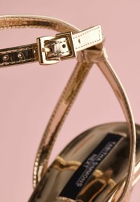 Guld sandal med stropper og en skinnende metallisk finish, med justerbare ankleremme og en glat, rundet tå. Detalje af spænde ses.