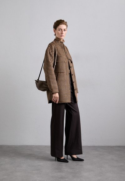 Bruine geruite oversized jas, wijde zwarte broek, zwarte puntige hakken en een kleine bruine schoudertas. Glad materiaal met contrasterende patronen.