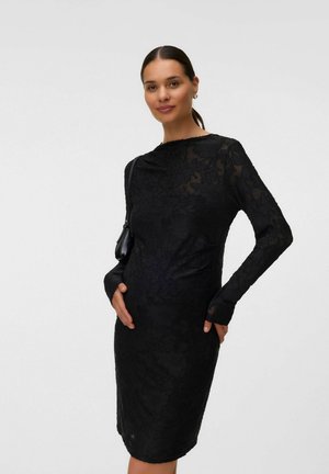 Mujer con vestido negro de manga larga con encaje floral sosteniendo un pequeño bolso negro de hombro con la mano apoyada en su vientre, de pie sobre un fondo blanco.