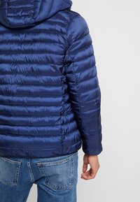 Donkerblauwe pufferjas met horizontaal doorgestikt ontwerp, voorzien van een capuchon. Het buitenmateriaal oogt glanzend en glad en biedt isolatie.