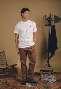 Weißes kurzärmeliges T-Shirt mit kleinem braunen Elefantenemblem, kombiniert mit braunen Hosen und schwarzen Schuhen. Der Hintergrund zeigt einen Garderobenständer und einen Stuhl.