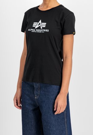 Femme portant un t-shirt noir Alpha Industries avec un logo blanc et un jean bleu foncé, debout devant un fond clair uni.