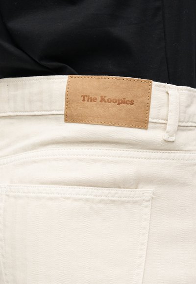 Κοντινή λήψη λευκού τζιν παντελονιού με μια ταμπέλα από καφέ δέρμα ραμμένη πάνω από την πίσω τσέπη που γράφει "The Kooples."