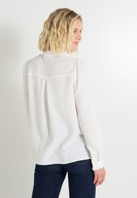 Camisa blanca de manga larga texturizada con detalle plisado en la parte posterior y puños abotonados, combinada con jeans oscuros.
