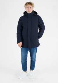 Mørkeblå polstret jakke med høj krave og hætte, der har en frontlynlås og trykknaplukninger, parret med lyseblå jeans og hvide sneakers.