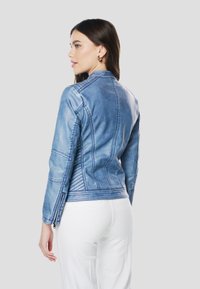 Blauwe spijkerjas met een getailleerd ontwerp, voorzien van een lichte wassing, gestructureerde naden en subtiele versieringen bij de schouder en manchetten.