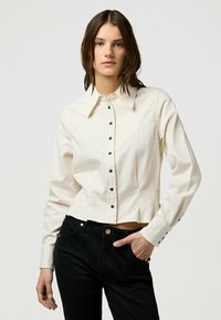 Camicia bianca corta con bottoni neri e dettagli a yoke in stile western, abbinata a pantaloni neri con accenti in rame.
