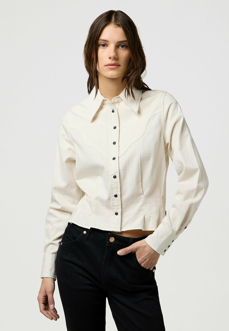 Camicia bianca corta con bottoni neri e dettagli a yoke in stile western, abbinata a pantaloni neri con accenti in rame.