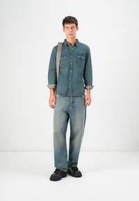Dunkelblauses Jeanshemd mit zwei Brusttaschen und Knopfverschluss, kombiniert mit hellblauen, locker sitzenden Jeans und schwarzen klobigen Schuhen.