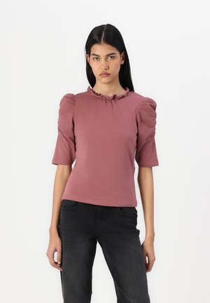 Un haut rose poussiéreux à manches courtes avec un col rufflé et des épaules bouffantes, fabriqué à partir d'un tissu doux et texturé. Associé à un jean noir.