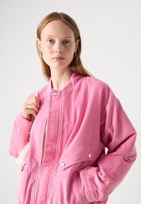 Rosa corduroy bomberjakke med ribbet tekstur, glidelås-lukking, sidelommer og trykknappdetaljer. Modell som holder en veske.