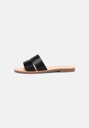 Flad sandal med tanfarvet sål og to sorte remme, en glat og en tekstureret, sat mod en ensfarvet grå baggrund.