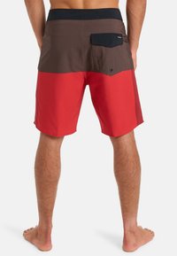 Homme debout pieds nus portant un short coloré en rouge et marron avec une taille noire et une poche arrière avec rabat.
