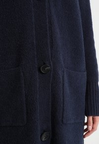 Námořnický vlněný kardigan s texturovaným povrchem, dvěma předními kapsami, černými knoflíky a žebrovanými manžetami pro volný střih.