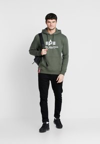 Olijfgroen hoodie met wit logo, voorzak en trekkoord. Zwarte broek en zwarte sneakers. Minimalistisch ontwerp op een eenvoudige achtergrond.