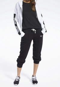 Sudadera blanca con cremallera y logotipo negro, camiseta negra y pantalones jogger negros con cordón, combinados con zapatillas deportivas negras con cordones blancos.