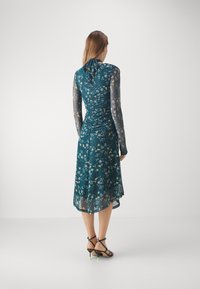 Vestito floreale turchese con maniche lunghe trasparenti, schiena arricciata e orlo fluente. Sandali neri con dettagli al tacco verde neon.