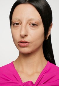 Nærbilde av et ansikt med rett svart hår, iført en fuchsia topp med knutedetalj. Kjennetegn inkluderer en leppepiercing og glatt hud.