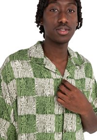 Camisa de cuadros verde y blanca de manga corta con un patrón de vetas de madera, que presenta un collar y un único bolsillo en el pecho.