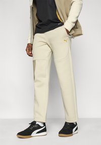 Μπεζ φόρμα με πλαϊνές τσέπες, που διαθέτει ένα ανοιχτό χακί πάνελ και κίτρινο λογότυπο Puma. Φοριέται με μαύρα αθλητικά παπούτσια και γκρι-μαύρο πουκάμισο.