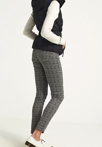 Kobieta ubrana w czarną kamizelkę puchową, szarą bluzkę z długim rękawem, wzorzyste szare spodnie o fakturze oraz białe buty na sznurówki stoi bokiem.