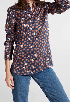 Kvinde iført en mørkeblå skjorte med orange og hvide polkaprikker og blå jeans, poserer med den ene arm hævet.
