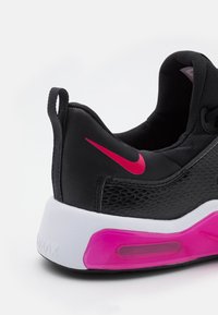 Zapatilla deportiva negra con textura de malla, acentos en rosa y una suela blanca. Presenta un lazo en el talón para facilitar el calzado.