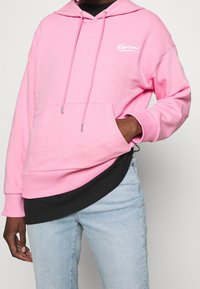 Felpa rosa in cotone con cappuccio, stringa, tasca frontale e logo bianco. Indossata sopra una maglia nera, abbinata a jeans azzurri chiari.
