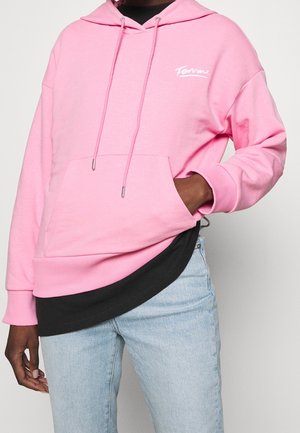 Felpa rosa in cotone con cappuccio, stringa, tasca frontale e logo bianco. Indossata sopra una maglia nera, abbinata a jeans azzurri chiari.