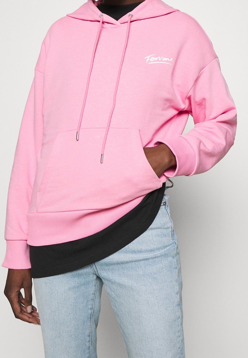 Felpa rosa in cotone con cappuccio, stringa, tasca frontale e logo bianco. Indossata sopra una maglia nera, abbinata a jeans azzurri chiari.