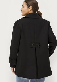 Zwarte wollen jas met een brede kraag, zichtbare stiksels, twee grote knopen aan de achterkant en omgeslagen manchetten. Lichtjes oversized pasvorm.